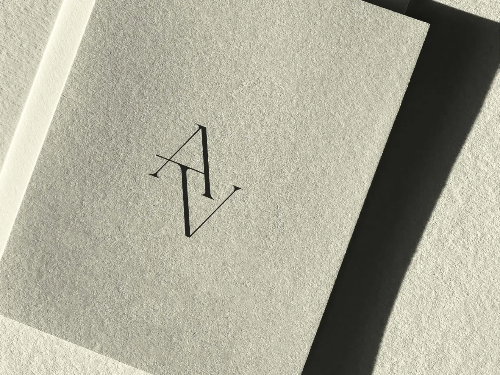 Black serif AV monogram printed on a textured sage-grey card in hard sunlight and shadow, for Aesthevision.