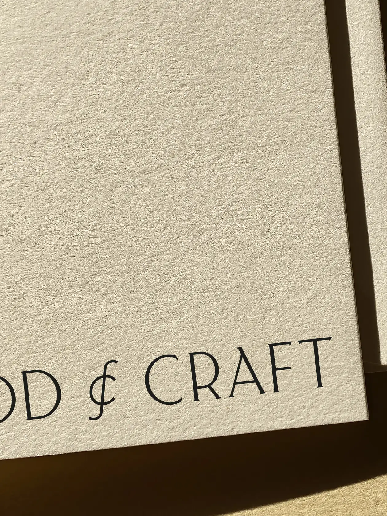 Close-up of a textured cream card corner printed with a black serif wordmark in warm sunlight, for Food & Craft.