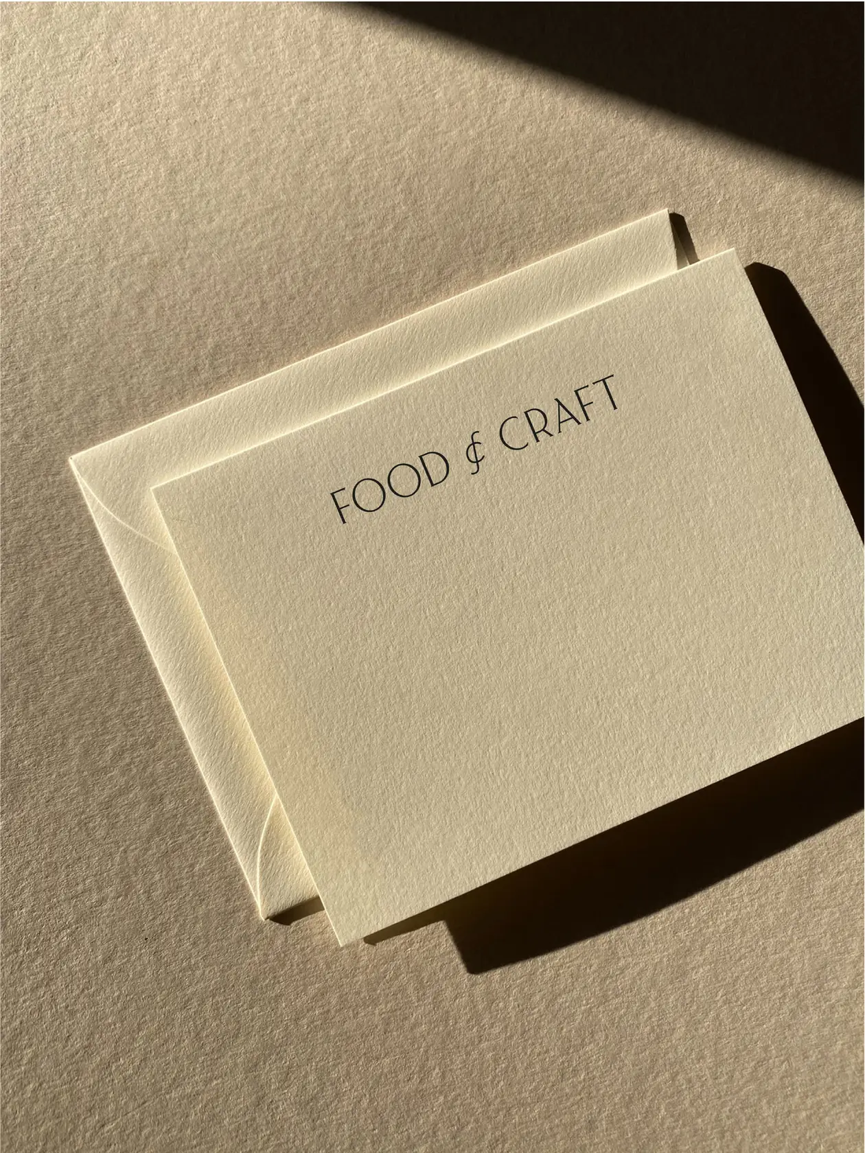 Cream note card printed with the Food & Craft wordmark on a matching envelope in raking sunlight, for Food & Craft.