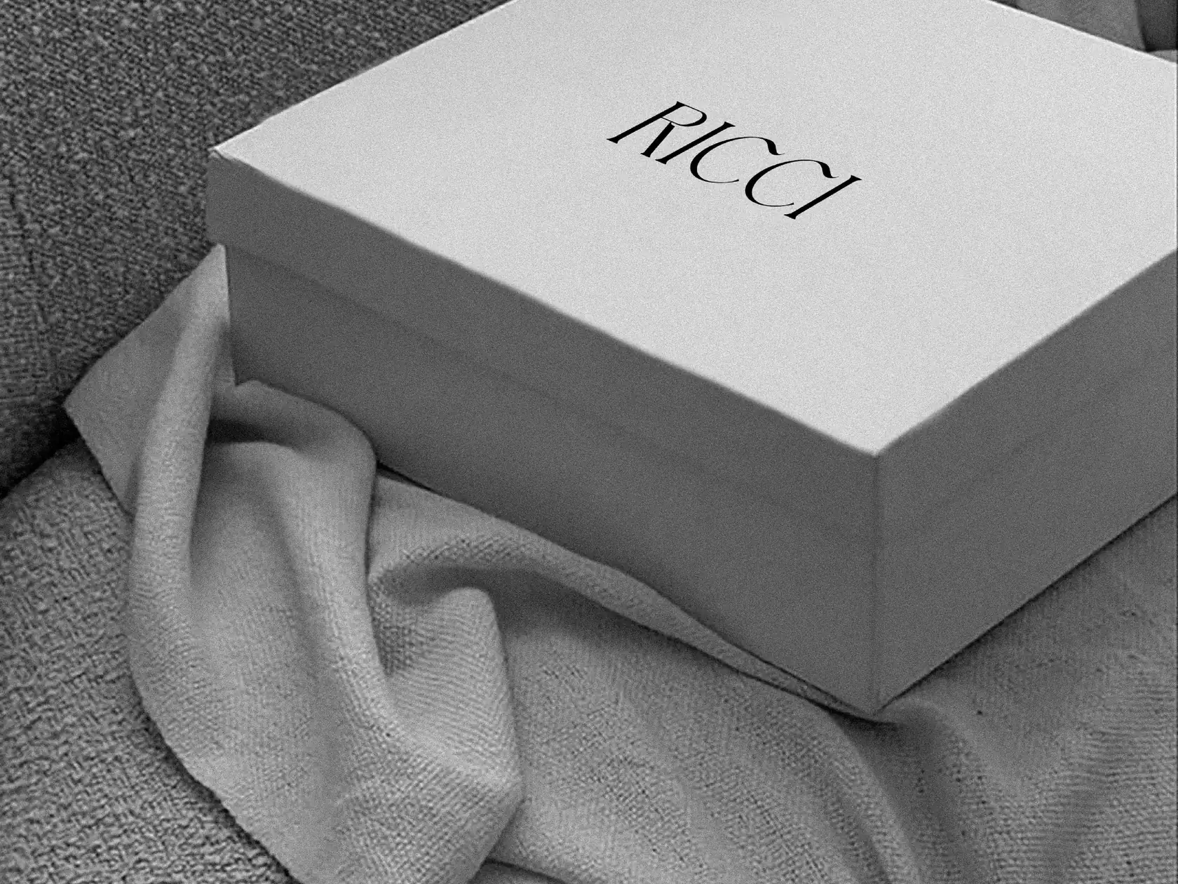 White rigid gift box with a black RICCI wordmark on the lid, resting on a draped grey knit throw, for Ricci.