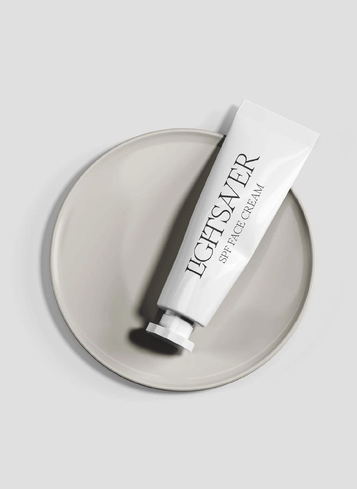 White SPF face cream tube with black serif lettering resting on a pale grey ceramic plate, for Lightsaver.