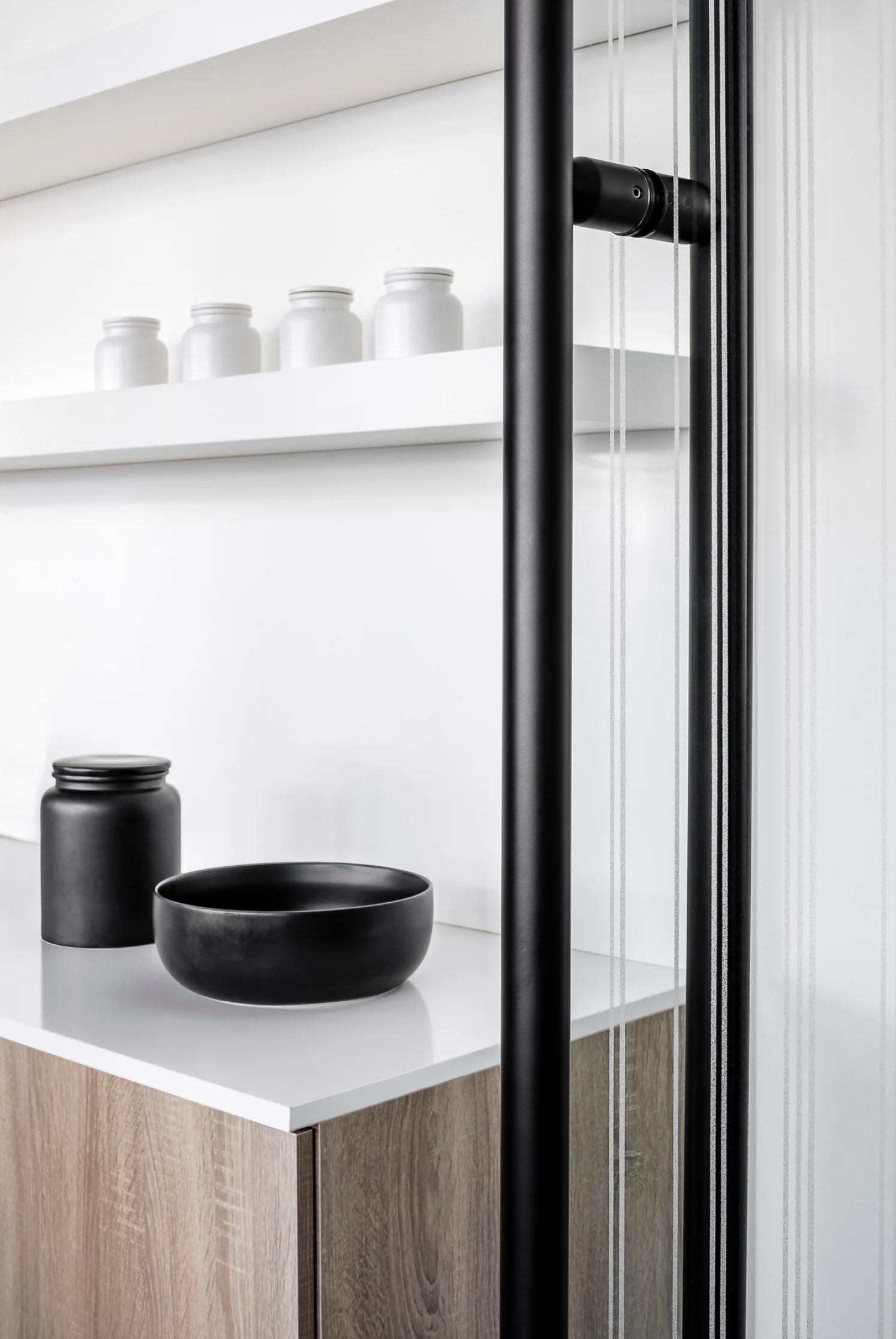 Black glass door handle beside a white counter with black ceramic jar and bowl, white jars on a shelf, for Sire Design.