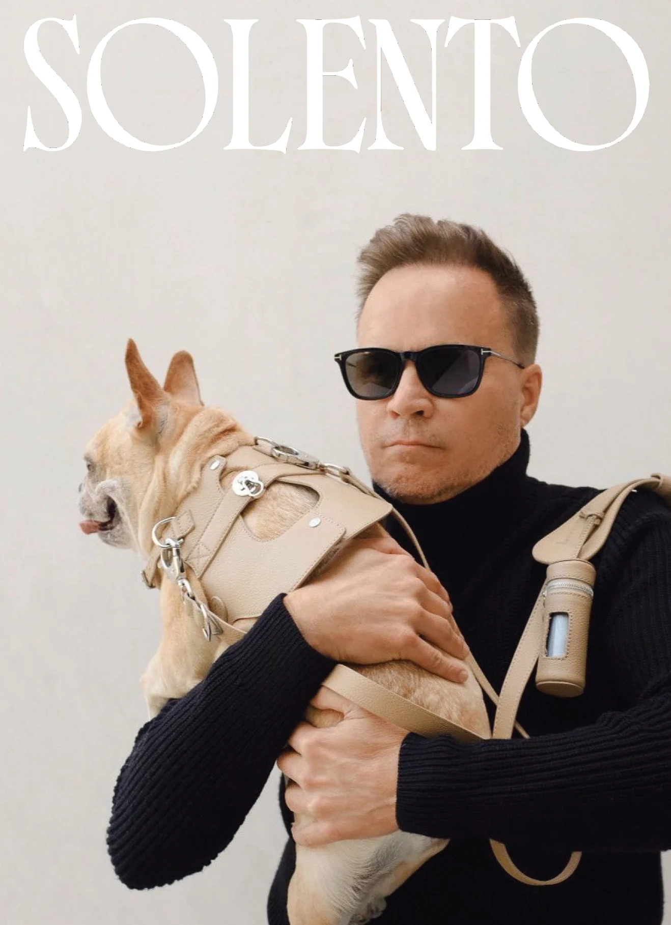 Man in sunglasses and black turtleneck holding a French bulldog in a beige leather harness under the Solento wordmark, for Solento.