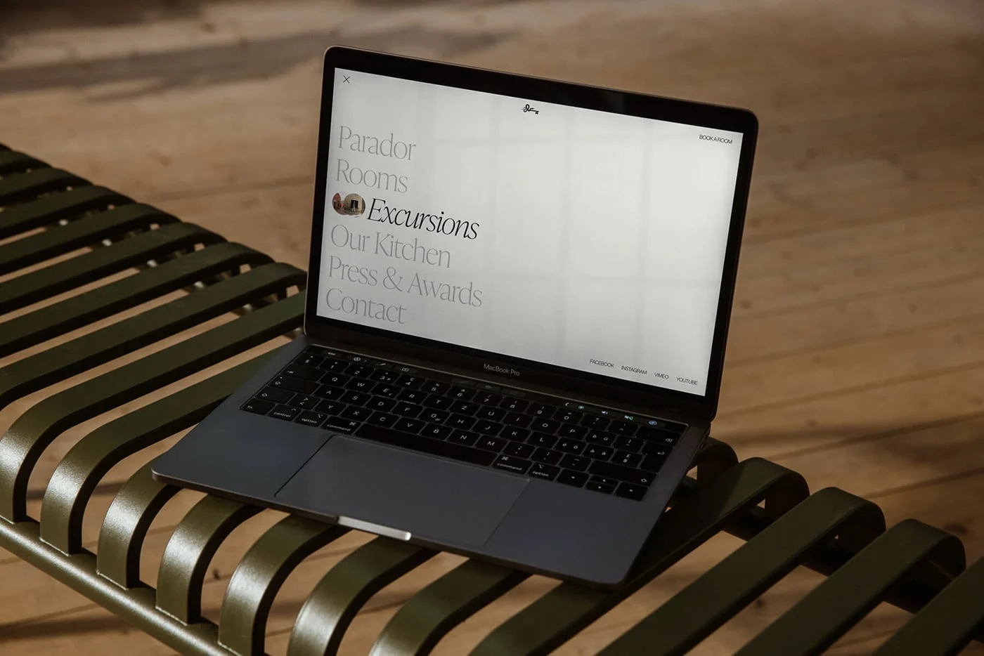 Laptop on a green slatted bench showing a website menu with Parador, Rooms, Excursions and Our Kitchen links, for Cirqa.